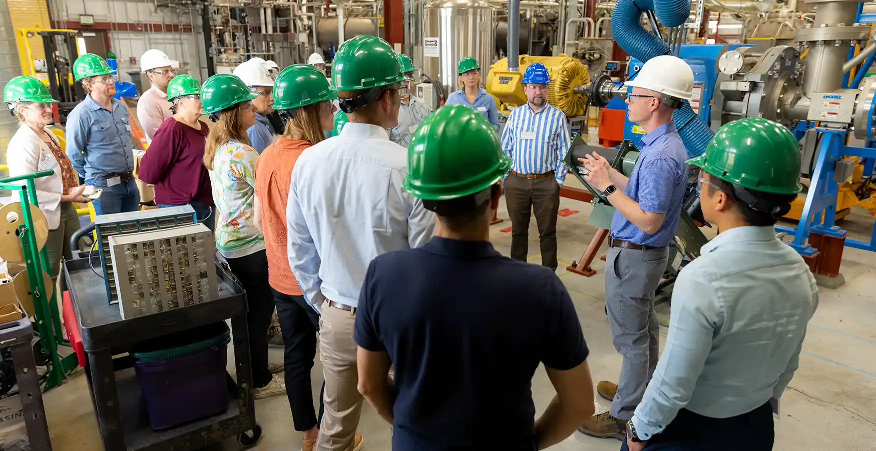 A group of people wearing hard hats in a laboratory listen to a tour guide speaking.