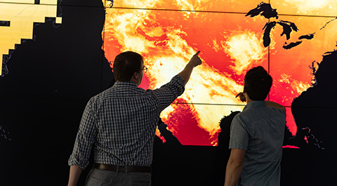 Two people point at heat map data on large display screen.