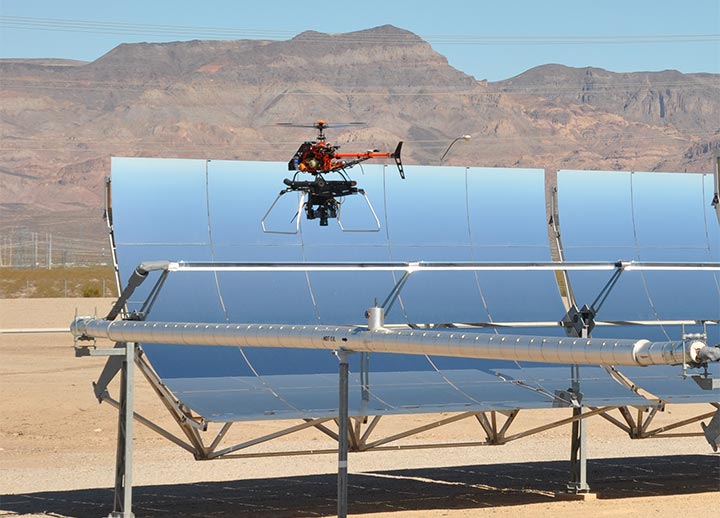 201909-facilities-23115 A small helicopter drone hovers near the top of a parabolic trough in the desert in front of mountains.