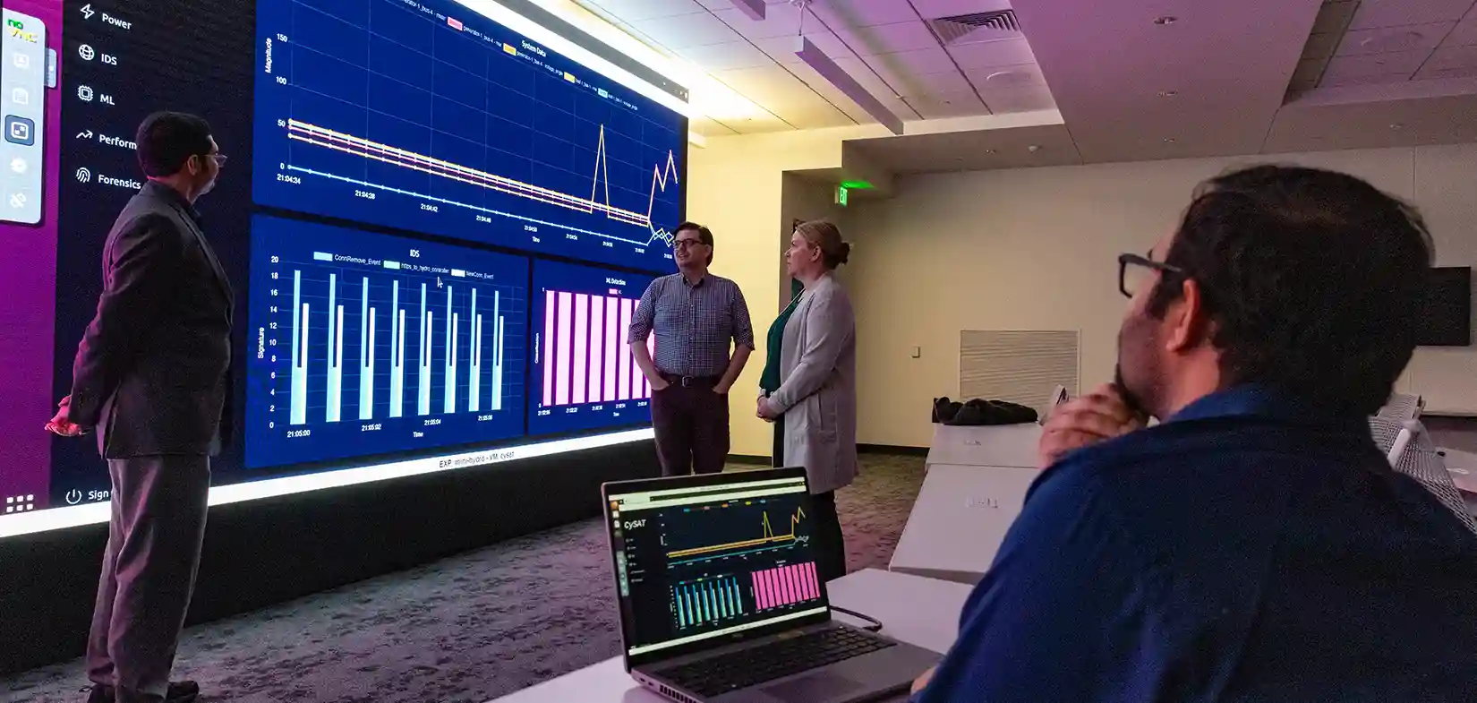 Researchers stand in front of large display screening discussing CySAT tool while another researcher sits in front of a laptop looking on.