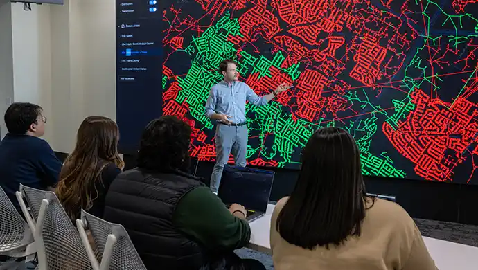 Person points to cyber data on large display screen in front of group of people.