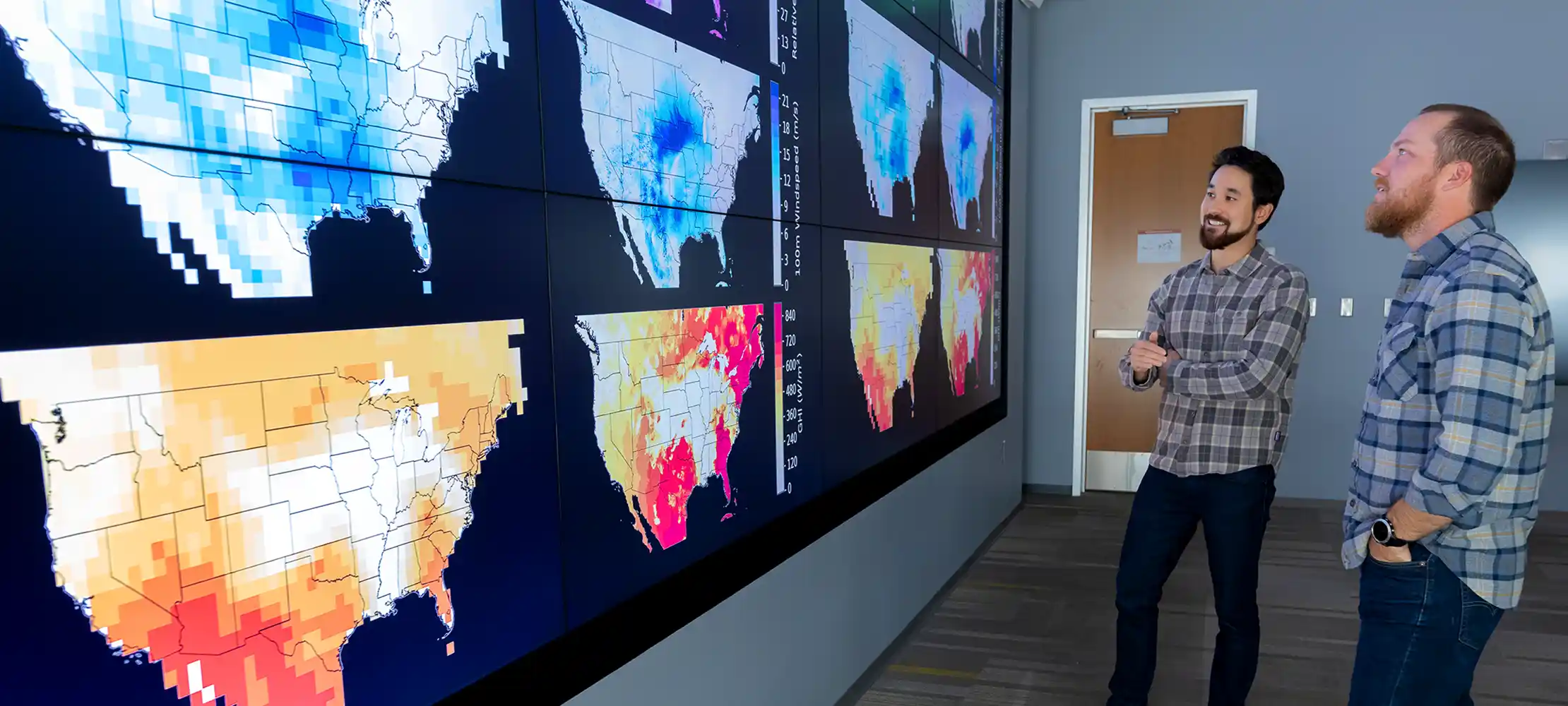Two researchers stand and look at temperature maps of the United States on a large display screen.