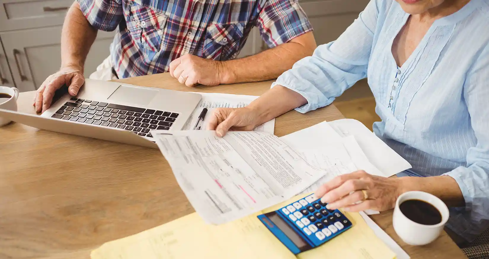 Two people look at a laptop and papers with a calculator and coffee mugs on the table.
