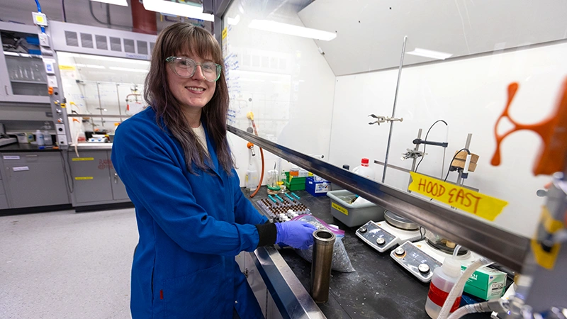 A person wearing protective eyewear and gloves and a lab coat stands beside a bench inside a laboratory containing various materials, including vials and scales.