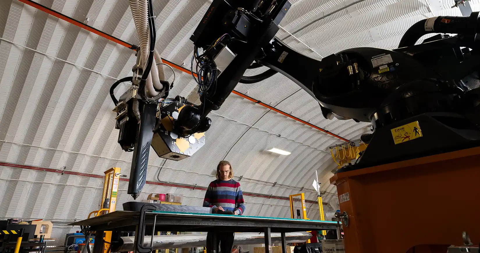 A person works at a table beneath a large industrial robotic arm inside a domed manufacturing facility.