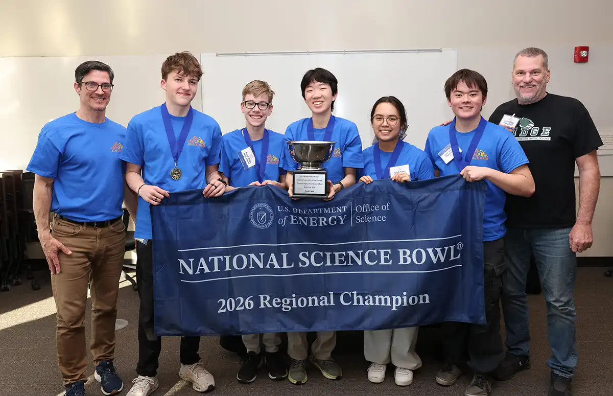 Seven people standing with a banner that reads National Science Bowl: 2026 Regional Champion. Five of the people are student competitors wearing medals, and the center student holds a trophy.