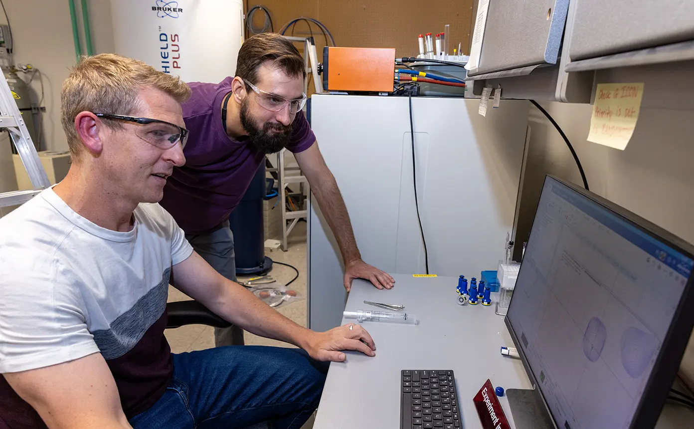 Two people in a lab look at data on a computer monitor.