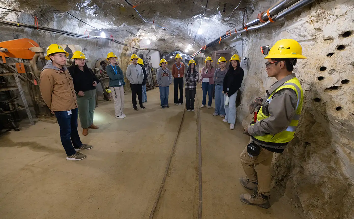 A group of students wearing yellow hard hats stands in an underground cave while an educator speaks.