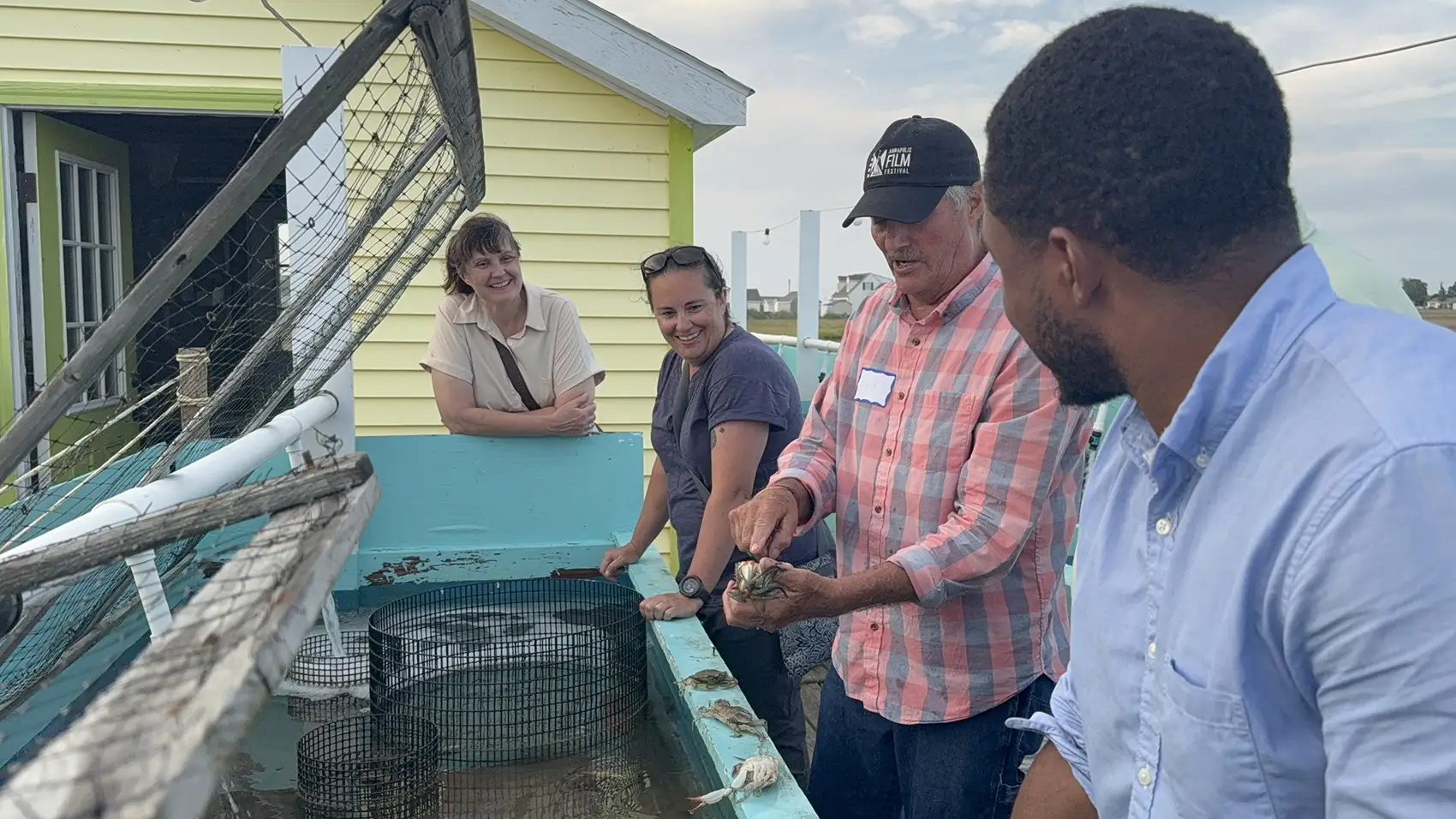 Three people standing next to crabbing equipment look at a man holding a small crab in his hand.