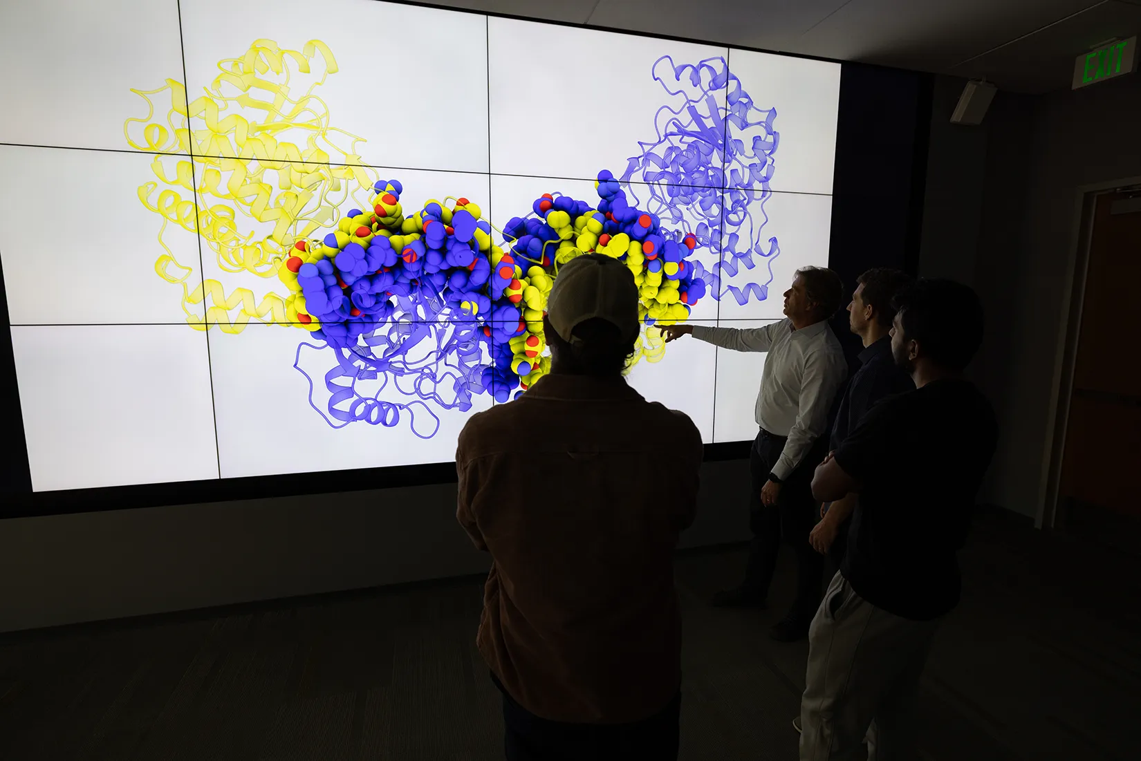 20260326-why-use-living-cells-nlr-is-making-chemicals-with-enzymes-alone-3d-visualization-102127 Four researchers examine a large visualization of an enzyme.