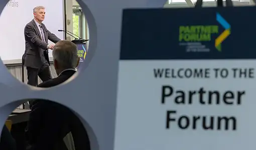 A person speaking at a podium with a sign with the text Welcome to the Partner Forum in the foreground.