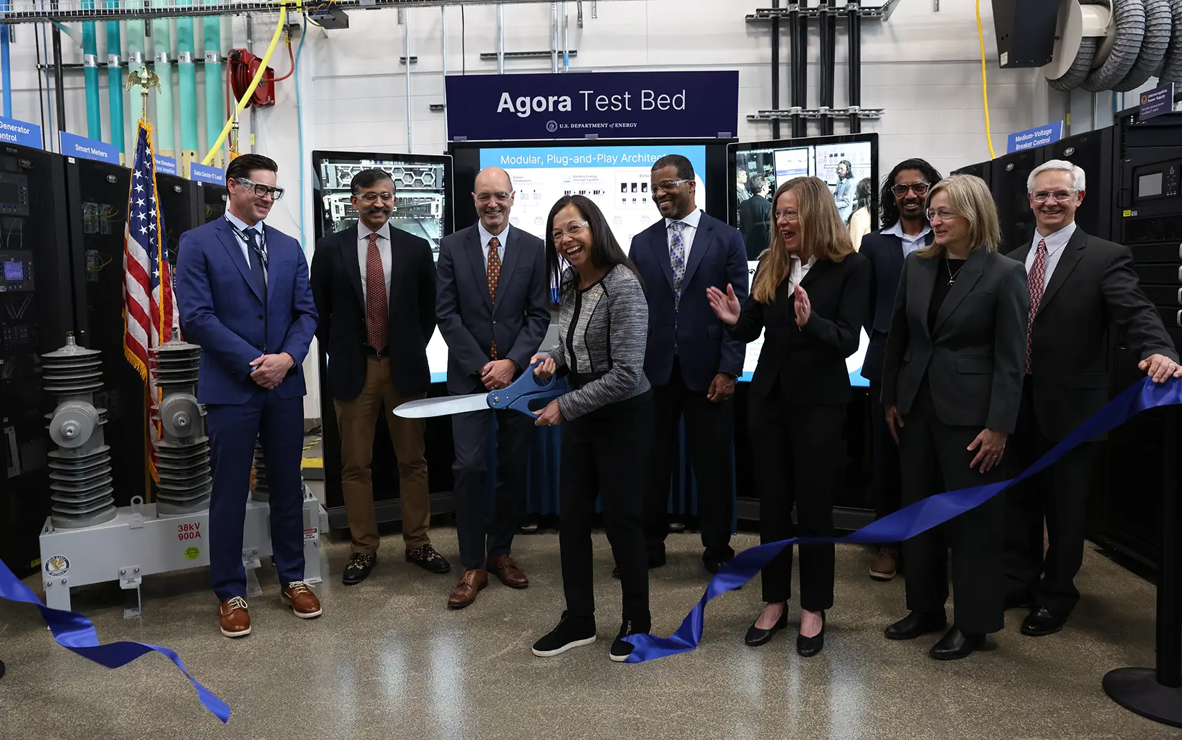 20260519-nlr-launches-agora-first-of-its-kind-large-load-grid-integration-test-bed-3787 Eight people smile as a person cuts a ribbon with giant scissors.