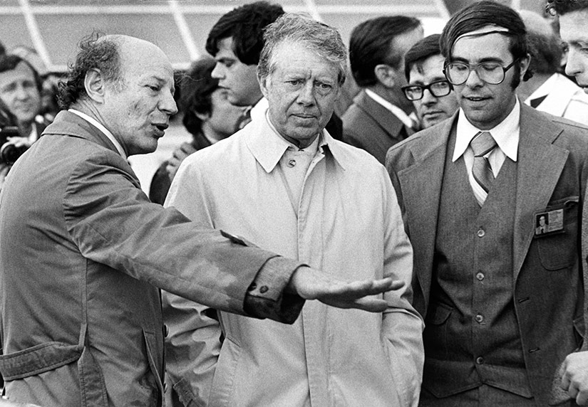 A black-and-white photo of people gathered around President Jimmy Carter outdoors with solar panels in the background.