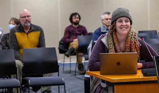 Continue reading about A woman sitting at a desk with a laptop and microphone to present while an audience behind her watches.