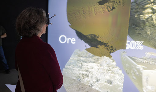 Woman wearing 3D glasses views a large data visualization screen showing mineral ore imagery and processing statistics.