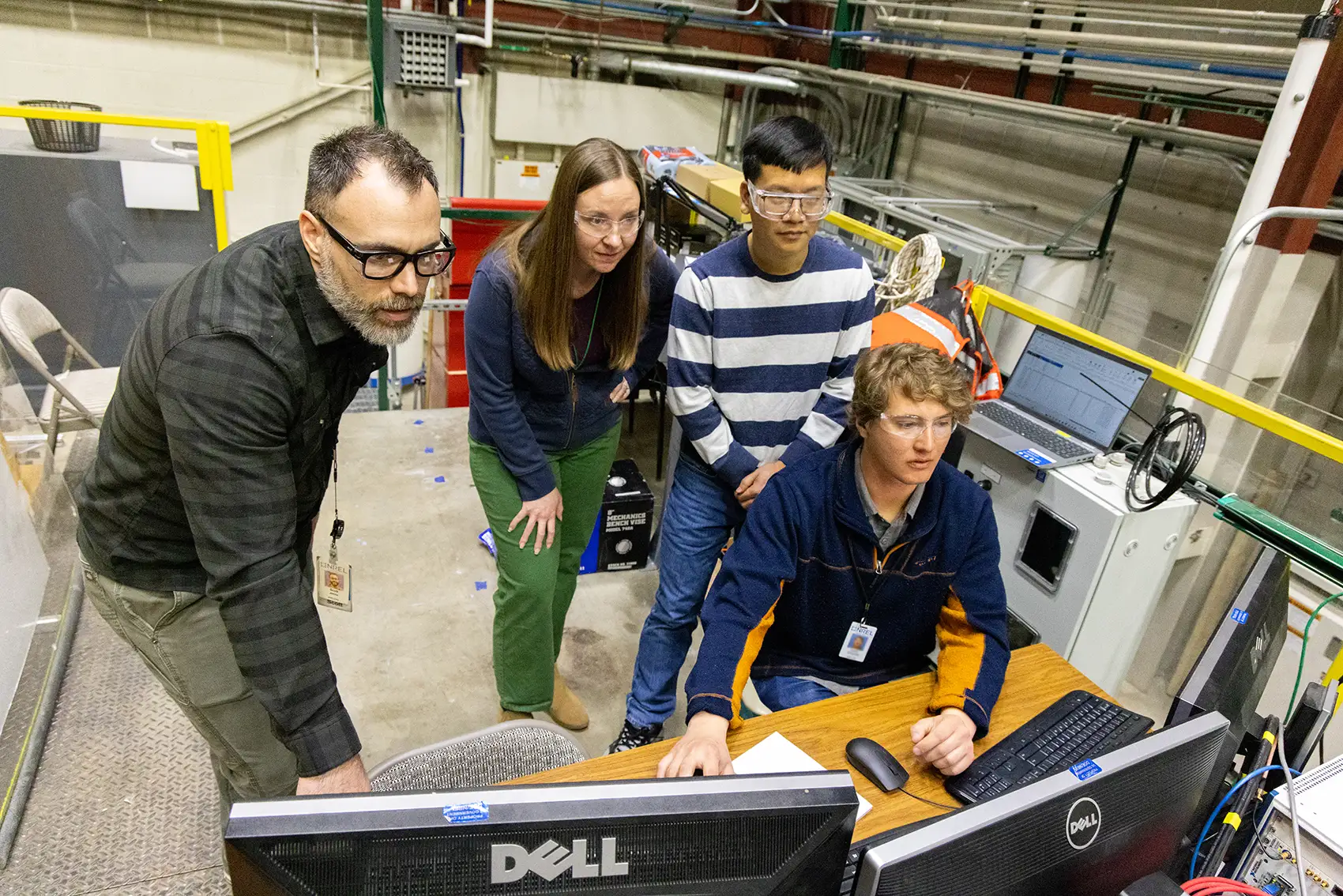 Four people stand or sit while looking at three computer monitors