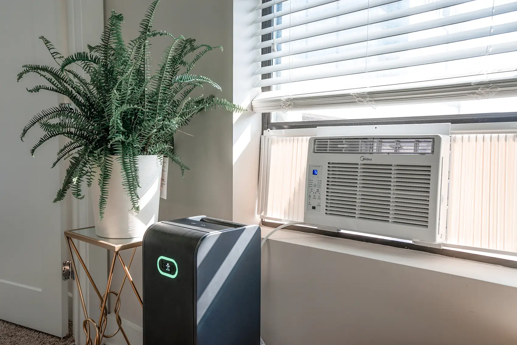 A black smart battery sits on the floor plugged into a window air conditioner beside a window and doorframe, with a plant on a nearby table against white walls and blinds.