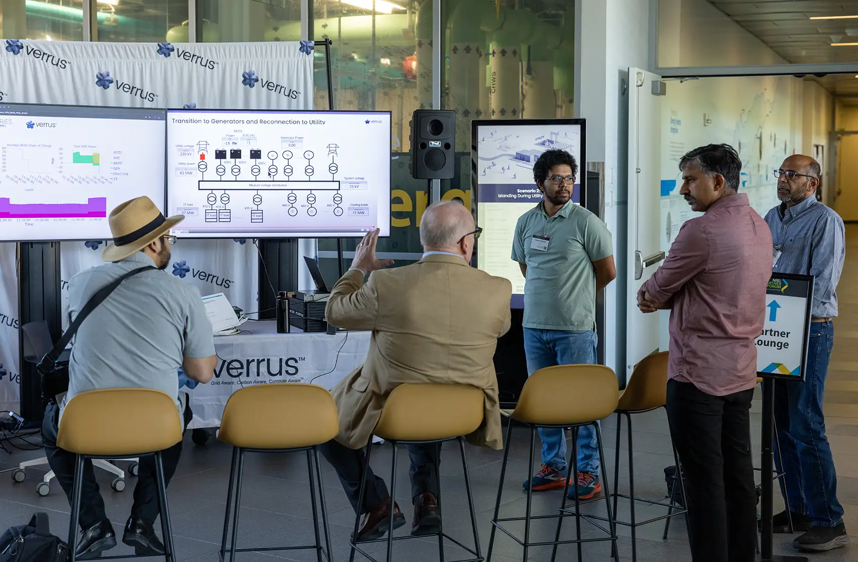 Five people conversing in front of a table reading “Verrus” and screens displaying data