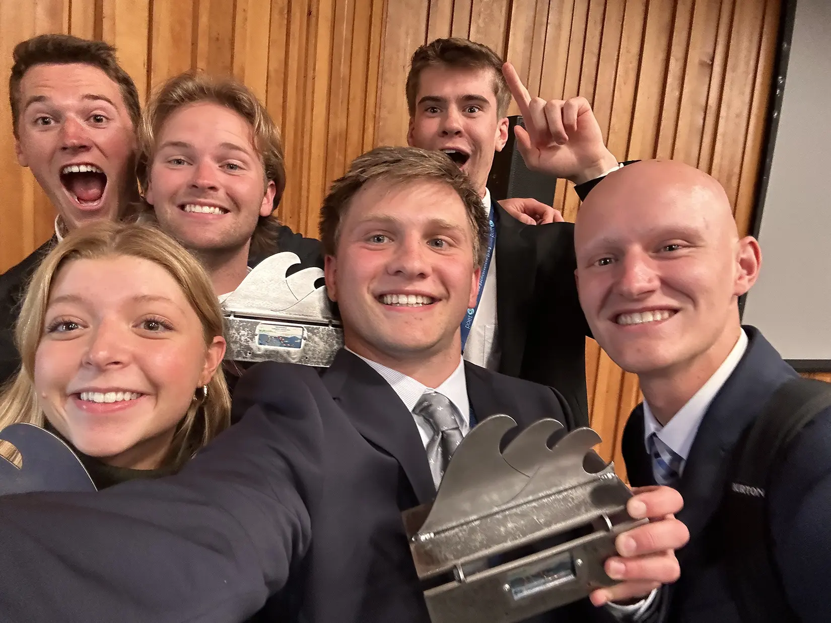 20260317-amped-to-shape-the-future-of-water-power-mecc-2024-winners A team of six smiling student competitors pose for a selfie. Three students are holding metal trophies featuring a wave design