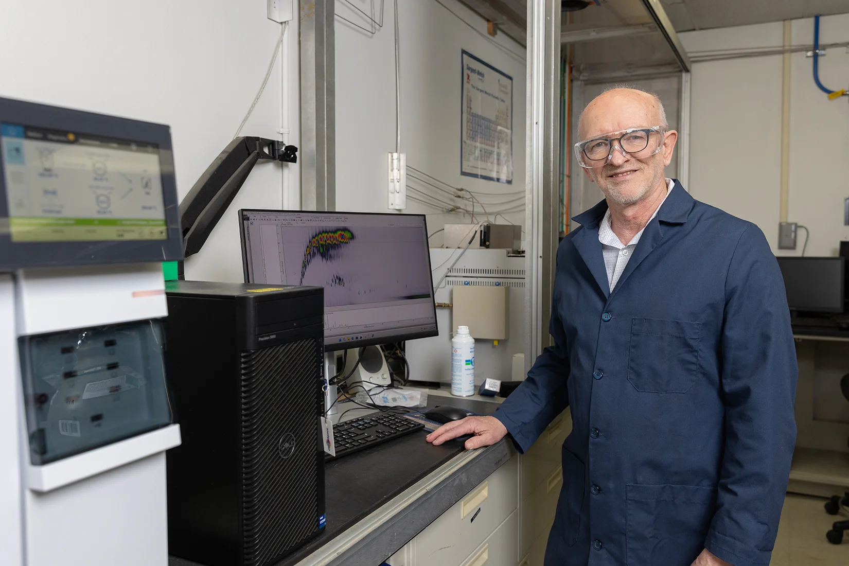A man wearing a dark blue lab coat stands at a desk with a computer and machine with another screen.
