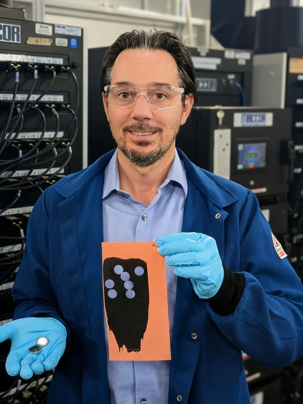 20260325-turning-biomass-into-graphite-could-help-secure-us-critical-mineral-supply-bertrand-tremolet-de-villers-graphite-electrode-batteries A researcher wearing blue gloves holds up a small graphite electrode and matching coin-sized batteries.