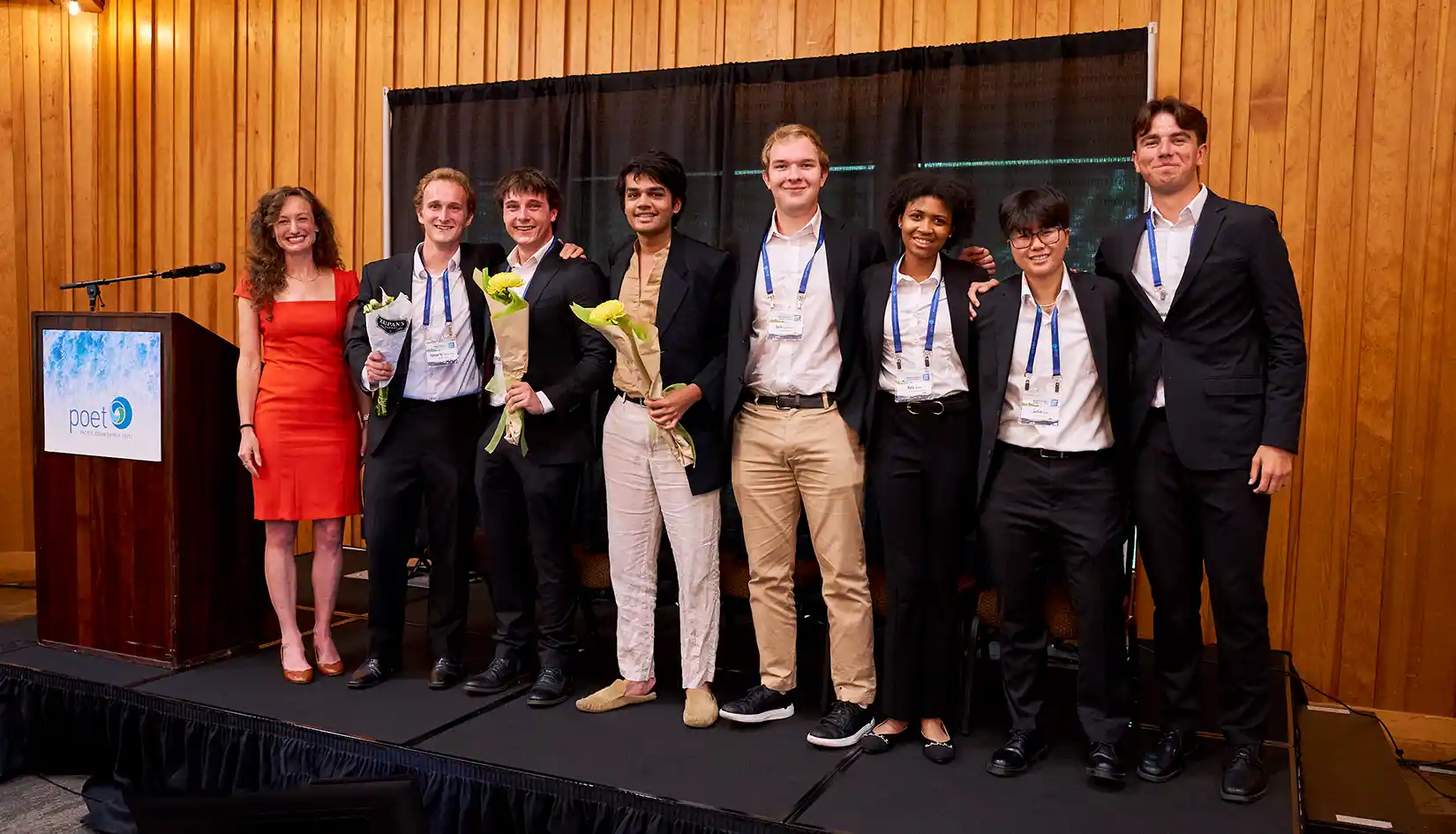 Photo of eight people, three of whom are holding bouquets, standing on a stage next to a lectern bearing the Pacific Ocean Energy Trust logo.