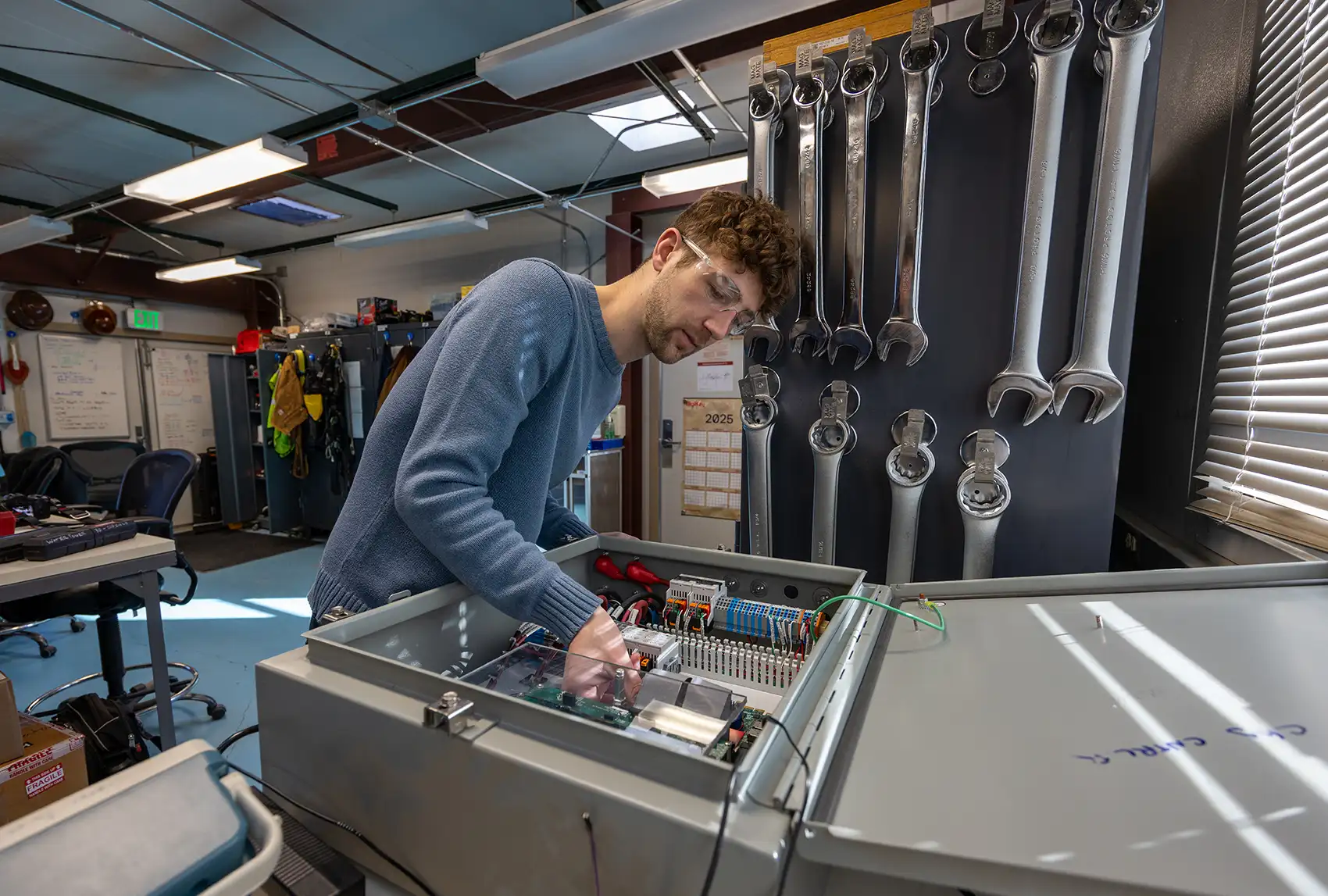 A young man wearing safety glasses and a blue sweater leans over a large, open gray electrical control box on a workbench.
