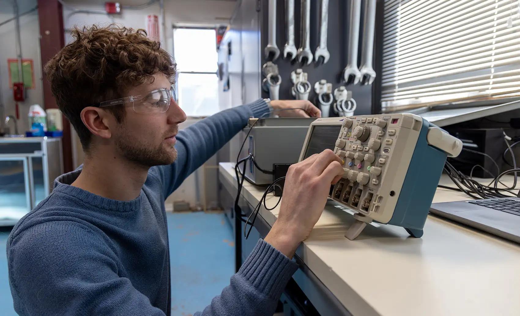 A person wearing safety glasses sitting at a workbench in a workshop, focusing intently as he adjusts the control knobs on a digital oscilloscope.