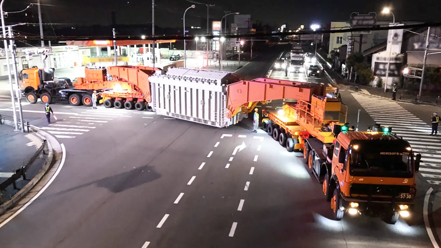 A nighttime photo of a massive heavy-transport configuration navigating an intersection. A large, rectangular white industrial load is suspended between two oversized, multi-axle orange transport rigs.