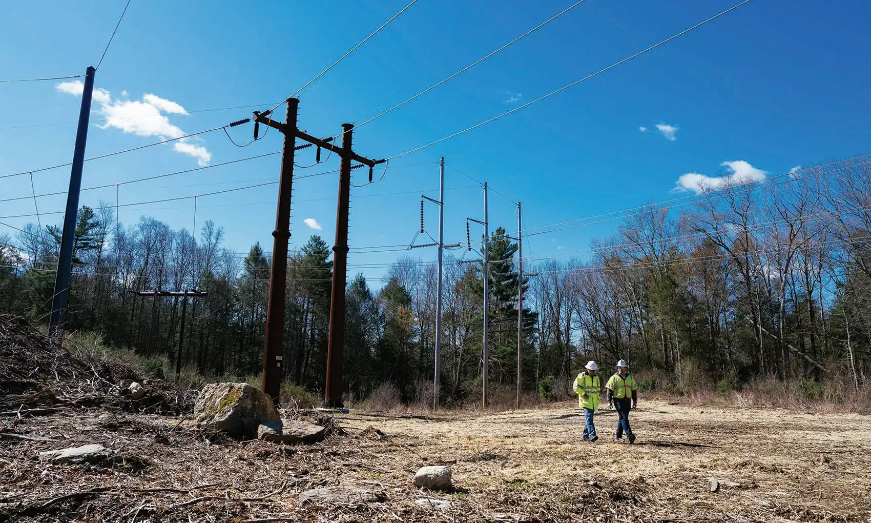 Two utility workers walk beneath high-voltage power lines in a cleared area on a sunny day.