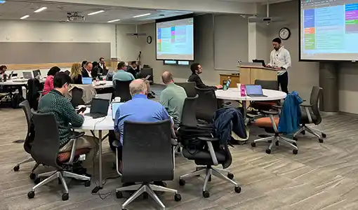 A group of people sitting around tables in a room while a person stands at a podium giving a presentation.