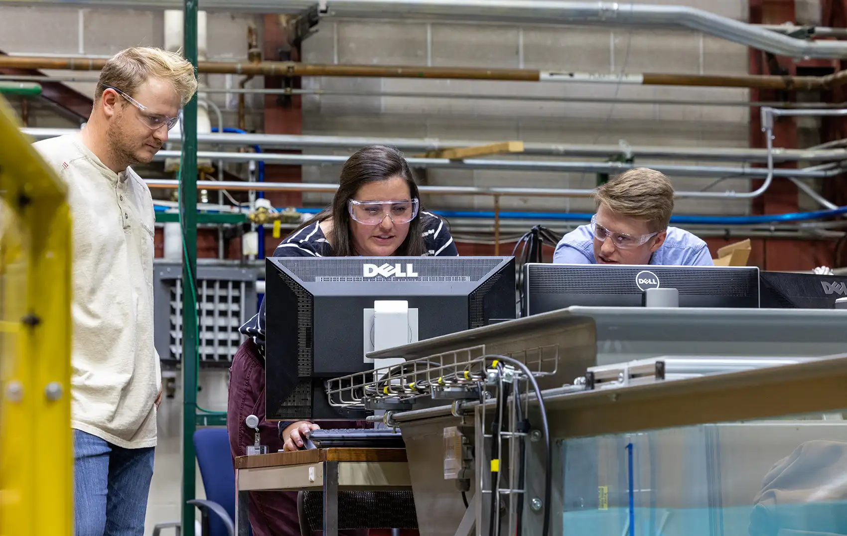 Photo of three people looking at a computer screen in a laboratory where three computer monitors are set up on a desktop next to a wave tank.