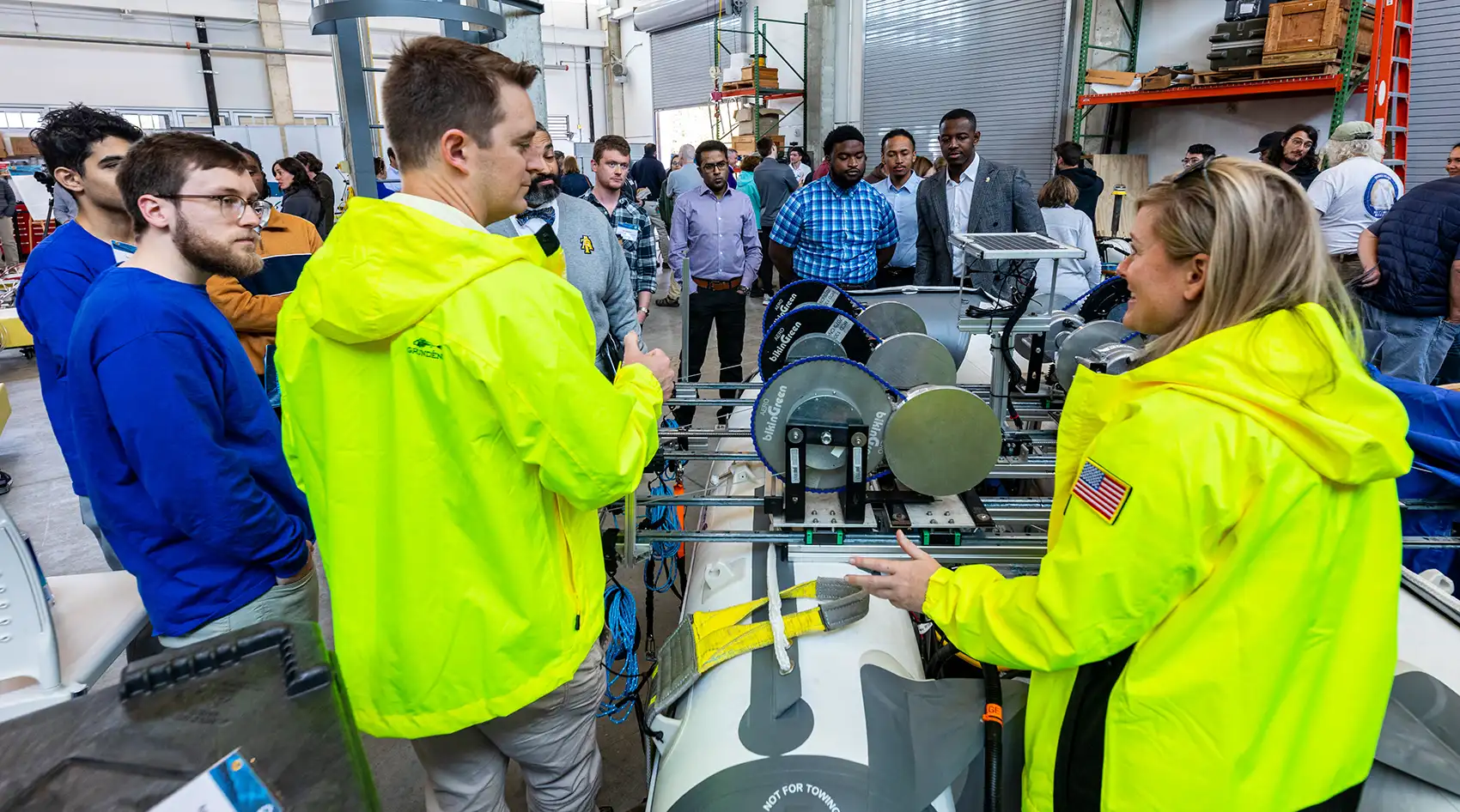 20261020-commercialization-hub-aims-to-ease-marine-energy-path-to-market-78063 Two people wearing yellow jackets speak to a group of people in a laboratory containing marine energy hardware.