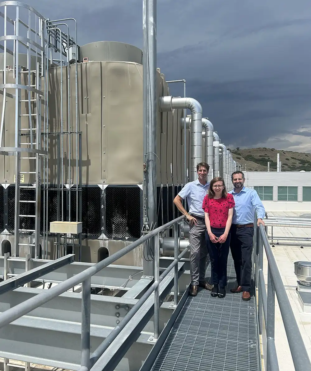 20261029-modeling-tool-can-help-developers-recycle-waste-heat-esif Three people stand on a suspended walkway next to a network of metal silos and pipes.