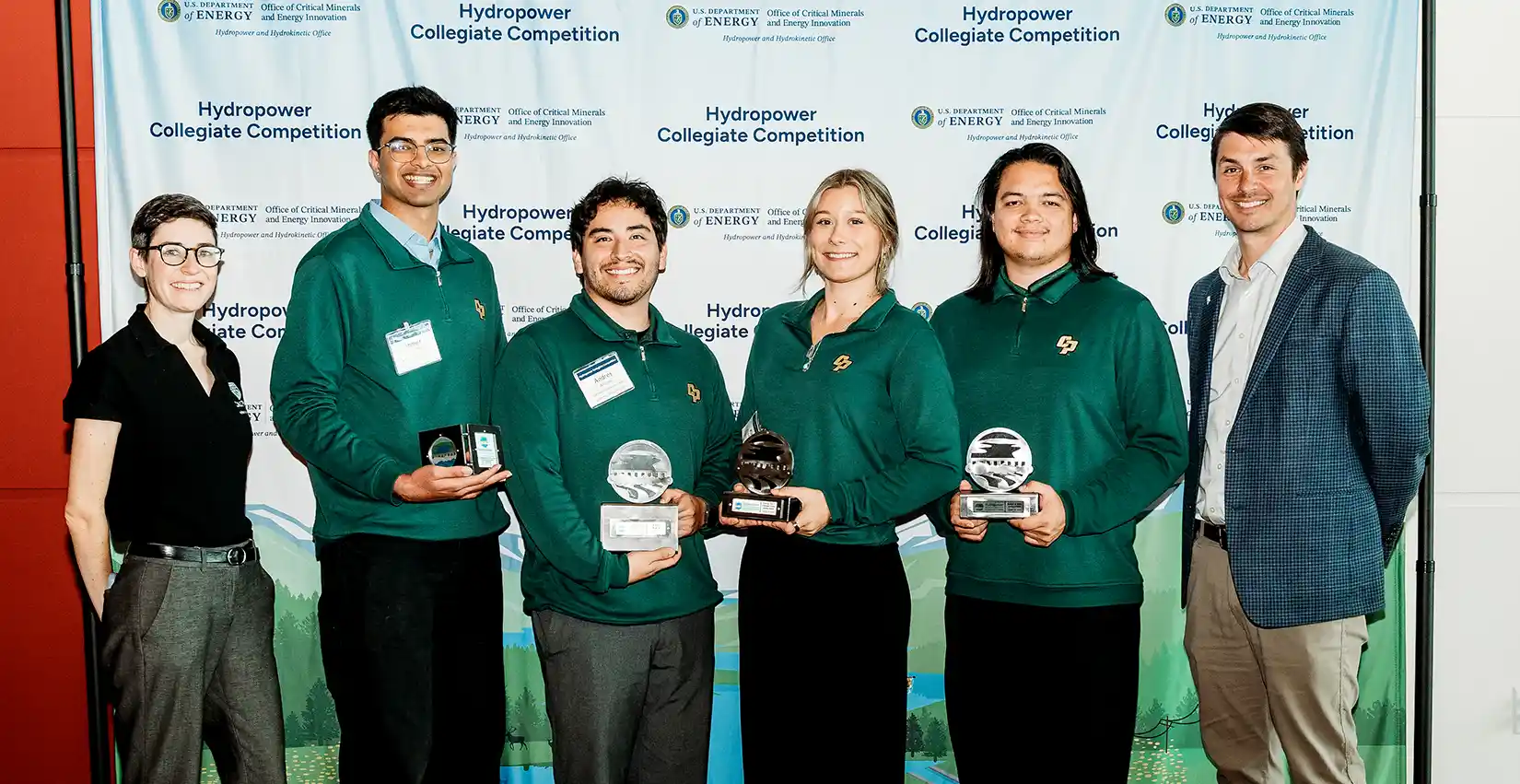 A photo of six people posing in front of a Hydropower Collegiate Competition sign. Four of the six are wearing green team jackets and holding awards in their hands.