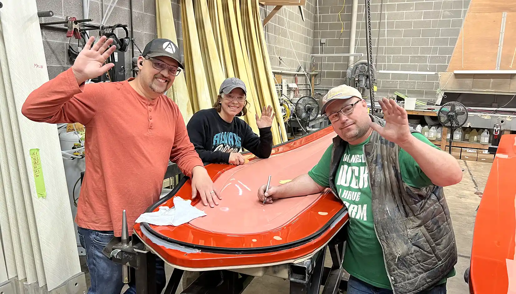 Three people smiling and waving at the camera as they work on a wind turbine blade in a laboratory.