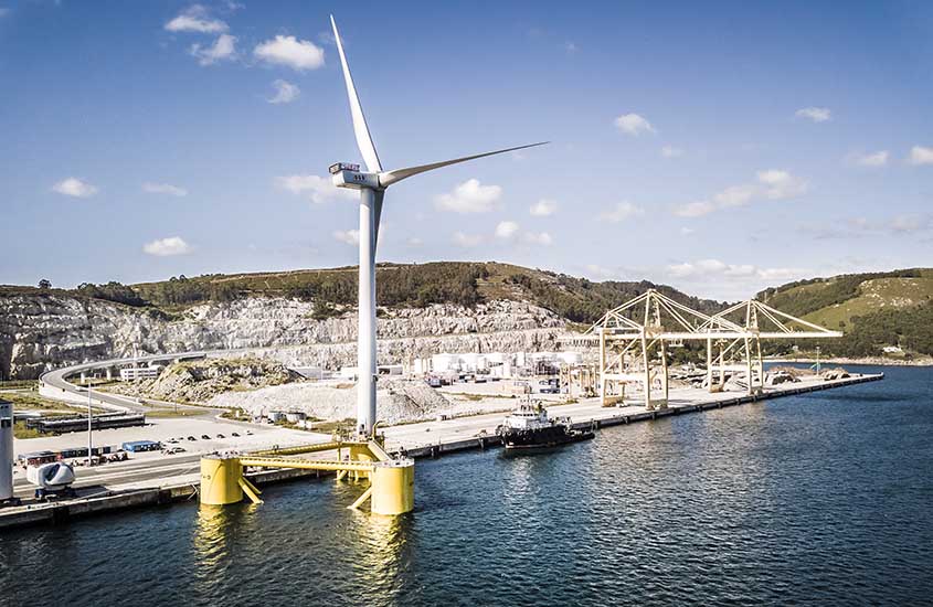 floating-spain-windfloat-atlantic-principle-power-dock90-center A wind turbine erected on one column of a three-sided structure floating in the ocean next to a dock.