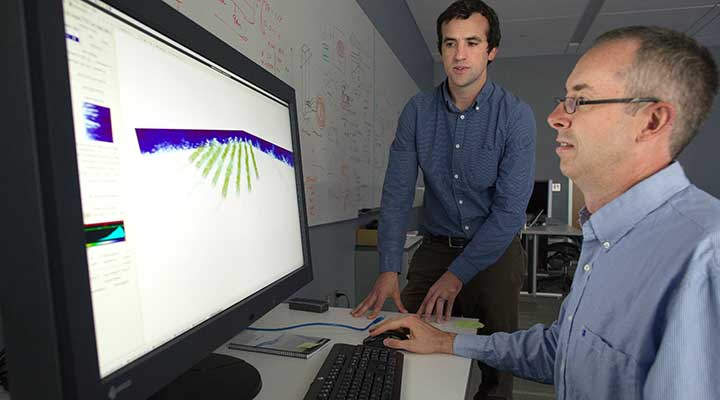 land-based-research-capability-tech-development A photo of two men looking at a computer monitor that displays blue and green wind-modeling graphics.